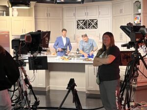 Television studio kitchen set with a chef preparing food at the counter while a male host reviews notes. Multiple large studio cameras and crew members are visible in the foreground.