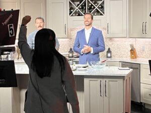 Chef Zac and host standing on a brightly lit TV kitchen set, smiling and preparing for a live segment while a crew member signals in front of the cameras.