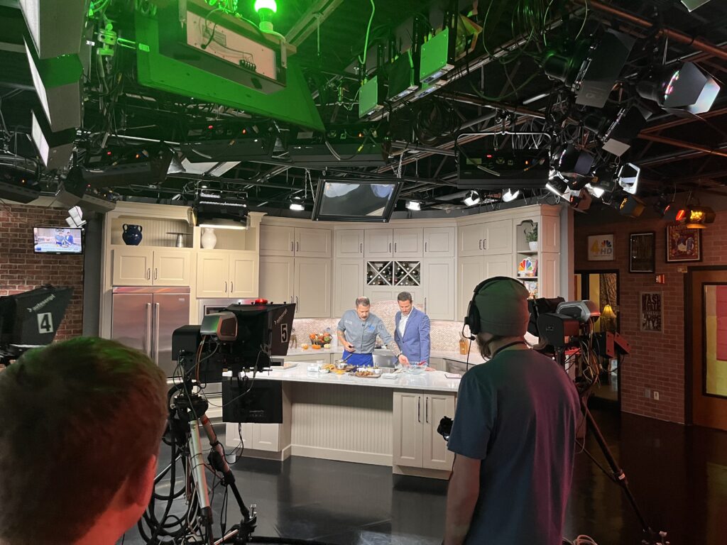 Wide view of a TV studio showing multiple cameras, overhead lighting, and crew members as a chef and host work together at a kitchen counter during a cooking segment.