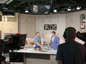 Chef Zac demonstrating a dish and speaking with the host on a TV kitchen set, filmed by several studio camera operators.